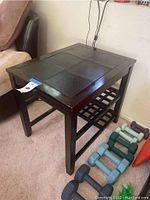 Front-side angle of the wooden end table showing inlaid leather panels on top, lower slatted shelf, and surrounding area with light weight dumbbells.