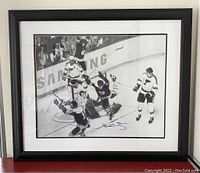 Framed black and white photo showing Bobby Orr celebrating a goal with opponents and fans visible. Samsung logo partially visible in the background on the boards.