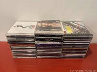 Stack of about 45 to 50 music CDs in jewel cases, stacked horizontally into three neat piles, showing various album covers and spines.