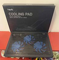 Cooling pad with three blue fans displayed in front of original Havit packaging box. The box is black with text indicating compatibility with laptops up to 17 inches.