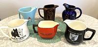 Full set of six vintage ceramic and porcelain Scotch whisky jugs displayed on a table, showing various colors and branding.