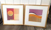 Two framed abstract lithographs by Amaina titled Sunset (left) and Sands (right) in wood frames shown leaning against a wall on the floor