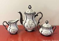 Front view showing coffee pot, creamer jug, and sugar bowl with lid, all decorated with sterling silver overlay on white porcelain.