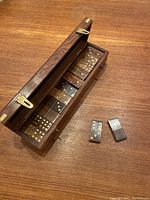 Open walnut domino box showing domino tiles with brass dots inside along with two tiles outside the box on a wooden surface.