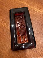 Oblique top view of signed Erik Höglund reddish-brown rectangular glass ashtray with abstract embossed geometric design recessed in center.