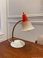 Photo of mid-century modern gooseneck desk lamp on a wooden table next to a chair, showing the lamp turned off with visible orange top shade and white flared bottom shade, metal base and flexible gooseneck arm.