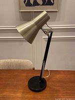 Full side view of black base with adjustable arm and brass-tone metal shade on wooden table with grey wall background.