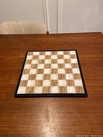 Chess board on a wooden table showing the full square grid with alternating brown and cream colored squares.