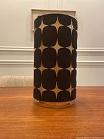 Front photo showing the black and dark brown geometric patterned cylindrical lamp shade with legs, standing on a wooden table.