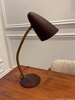 Vintage mid century bullet style desk lamp placed on wooden table showing full lamp profile with burgundy fibreglass bullet head and brass gooseneck.