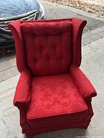 Front view of red vintage wingback chair with tufted back and floral damask pattern upholstery, outdoors next to a car.