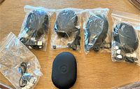 Five sealed plastic pouches containing individual pairs of earphones, USB cables, and ear tips arranged on a wood surface alongside a black silicone carrying case.