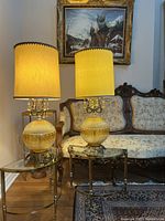 Pair of vintage brass base table lamps lit on side tables with a vintage upholstered sofa and a framed painting in the background.