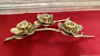 Side angle view of the brass branch and floral-shaped candle holder, showing the three arms and petal detailing in the candle cups on red surface.