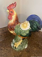 Front and side view of colorful ceramic rooster figurine with flower detail, sitting on brown surface.