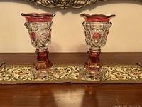 Pair of cranberry flash glass celery vases with intricate pressed patterns, displayed on a decorative table runner.