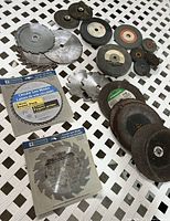 Overall view of lot showing many abrasive discs and circular saw blades in used and new condition on white lattice surface.