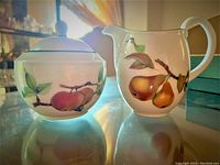 Photo displaying front view of sugar bowl with lid and creamer jug, showing fruit designs and overall aesthetic of fine porcelain pieces.
