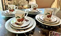 Four complete porcelain place settings displayed on a glass table, each with a cup, saucer, side plate, and dinner plate featuring the Evesham Vale pattern with orange fruit and green trim.