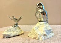 Two silver sculptures on white stone bases positioned on a textured surface against a plain background, showing overall scale and design.