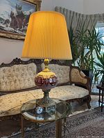 Overall view of the ruby glass table lamp with cream pleated fabric lampshade on glass and brass side table in living room setting.