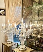 Two matching crystal candelabras placed on side tables in front of a window and display cabinet, both lit.