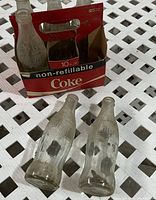 Photo showing six clear embossed Coca-Cola glass bottles in original red and black cardboard carrier sleeve marked 'non-refillable Coke', bottles empty and with wear.