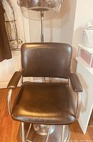 Front view of the black salon/barber chair showing padded vinyl seat and backrest with chrome arm supports and padded armrest tops.