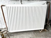 Photo showing one white Stelrad Softline Compact radiator with vertical ribs and original copper pipework attached, positioned against a wall on a concrete floor.