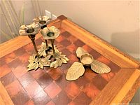 Photo of two vintage brass candle holders on a wooden table, showing one three-arm candle holder with a leaf base and flower-shaped cups alongside a single leaf-shaped candle holder.