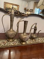 Three vintage Middle Eastern brass decanters arranged on a table runner showing ornate carving details and overall shapes.