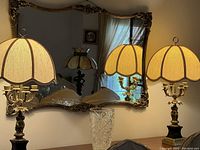 Pair of vintage Churb table lamps with cream fabric scalloped lamp shades and ornate bronze cherub candelabra bases, positioned in front of a decorative mirror and next to a cut glass vase.