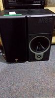 Two black B&W speakers shown side by side, one with front cover and one without, placed on carpeted floor.