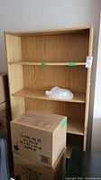 Front view of one wooden bookcase surrounded by boxes, showing 3 of the 5 shelves and light wood finish.