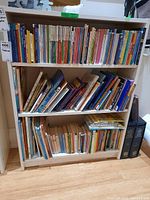 Three shelves on a white bookshelf full of various children's books, mostly paperback with colorful spines, arranged vertically and stacked horizontally in some areas.