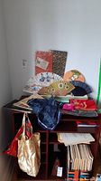 Overview photo displaying Asian art books stacked along bottom shelf, three fabric bags hanging, and a variety of hand fans arranged upright on the top shelf. Also shows packaged handmade paper sheets propped against the wall.