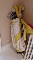 Set of right handed golf clubs in a white and yellow golf bag, standing upright against a beige wall and radiator, with yellow and white knit head covers visible.