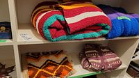 Two hand knit blankets - one multicolored striped blanket and one purple blanket, along with a ladies knit sweater folded on a shelf