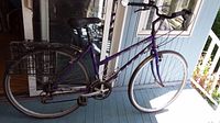 Side view of the purple Maxam ladies bicycle on a porch, showing frame, wheels, and rear basket.