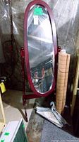 Full view of free standing oval mirror with wooden frame, showing adjustable angle supports and attached tag. Bottom area shows metal hangers on the floor and rolled bamboo blind leaning against wall.