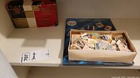 Photo showing a box filled with mixed loose postage stamps next to an unopened blue souvenir postage stamp album and a vintage red and beige box.