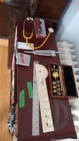 Photo showing a vintage yellow stethoscope, metal and wooden measuring rulers, and the wooden box containing the Sikes hydrometer set with brass weights.