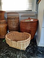 Photo showing the four wicker baskets arranged on a black marble floor near a window: a round basket with lid, a tall standing basket, a cylindrical basket, and an oval basket with handles.