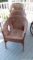 Four brown wicker chairs stacked on a wooden porch floor outside. Chair seats and backs are woven wicker with some visible wear and fading. Armrests curve around seat area.