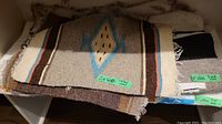 Stacked wool floor mats showing various geometric patterns and fringed edges with green tags labeling '2x wool place mat' and '1x wool floor mat'.