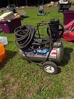 Side view of Campbell Hausfeld air compressor showing the cast iron tank, wheels, and attached coiled hose on grassy outdoor surface.
