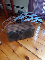 Bluetooth stereo speaker with two speaker cones and control knobs on front, black wood grain finish, placed on wooden floor next to a grey directional antenna with multiple elements and coaxial cable.