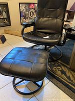 Full view of black leather-like chair with matching ottoman on tiled floor, showing padded seat, backrest and armrests with black circular metal base