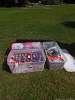 Three plastic storage totes on grass, filled with various holiday decorations including Christmas lights and ornaments.