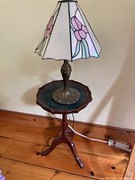 Side view showing wooden table with three legs and scalloped edge tabletop supporting stained glass lamp.